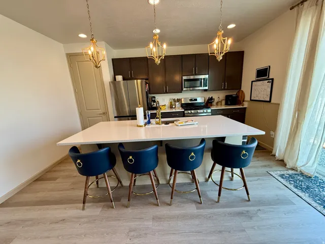 Open-concept dining and kitchen area featuring a white quartz countertop island, dark cabinetry with stainless steel appliances, wood-finish flooring, and three brass pendant lights suspended above the dining surface