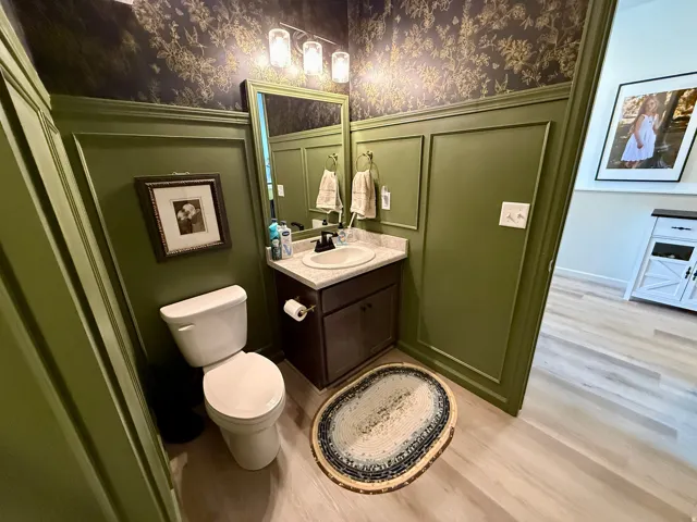 Elegant powder room featuring sage-green paneled walls with decorative upper wallcovering, dark vanity with integrated sink and countertop, framed mirror with sconce lighting, and wood-finish flooring