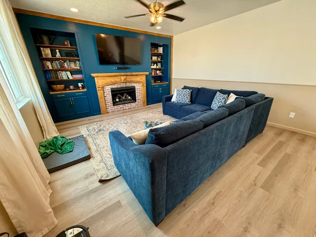 Built-in cabinetry with shelving, brick fireplace surround with wood mantel, wood-finish flooring, and a ceiling fan with integrated lighting