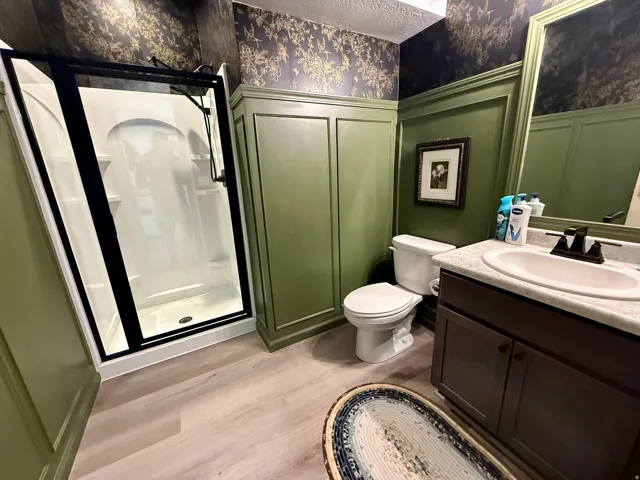Walk-in shower with black-framed enclosure, single vanity with integrated sink, wood-finish flooring, and green paneled wall accents