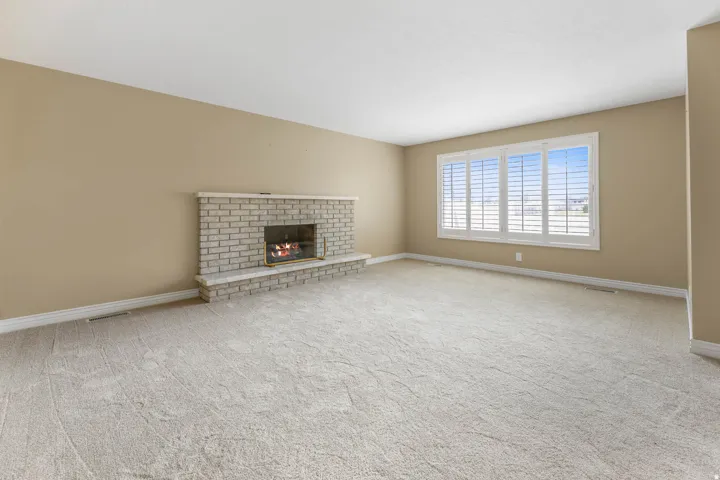 Neutral-toned carpeted flooring, a brick-surround fireplace with a stone mantel, and a window with white plantation shutters