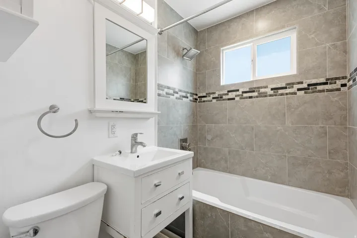 Bathroom featuring a white vanity with integrated sink, white framed mirror, tub with overhead shower, and floor-to-ceiling tile surround with decorative mosaic accent