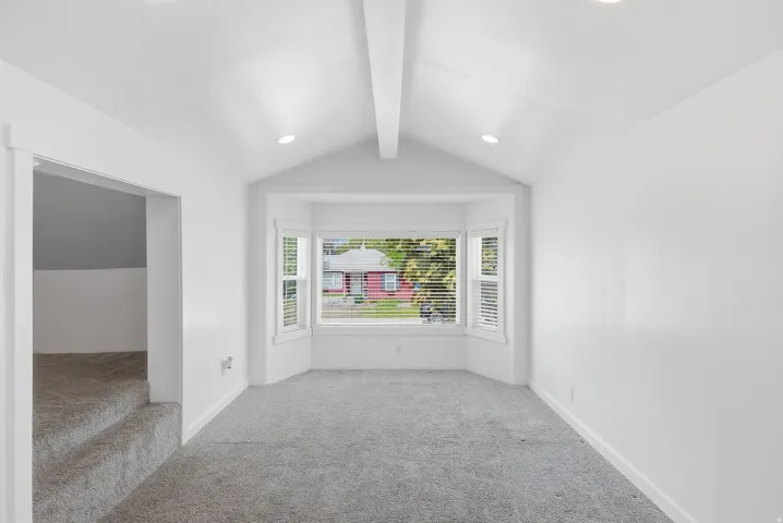 Vaulted ceiling with exposed beam, recessed lighting, bay window, light gray carpeting, and white wall finishes