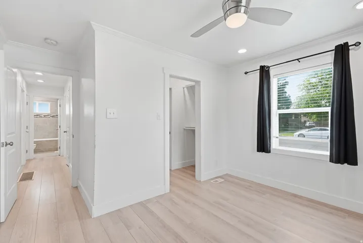 Bright bedroom with wood-finish flooring, large window with black curtain rod, ceiling fan with integrated lighting, and direct access to walk-in closet and ensuite bathroom
