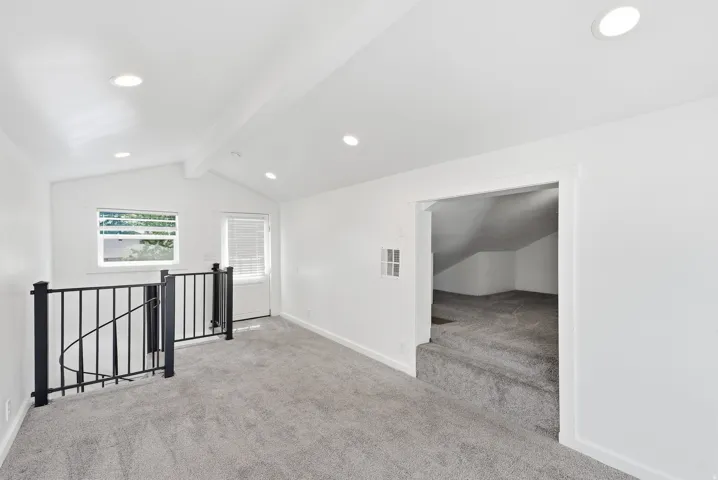 Vaulted ceiling with recessed lighting, neutral carpet, black metal railing, natural light from windows, and an additional carpeted area