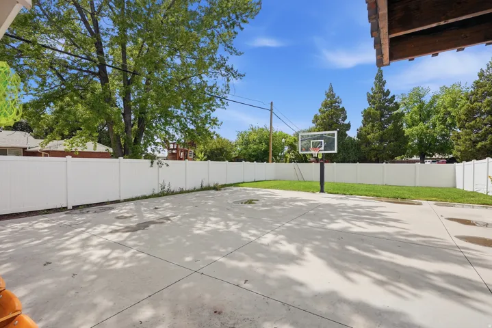 Expansive concrete patio with an attached basketball hoop, bordered by a white privacy fence and a manicured lawn