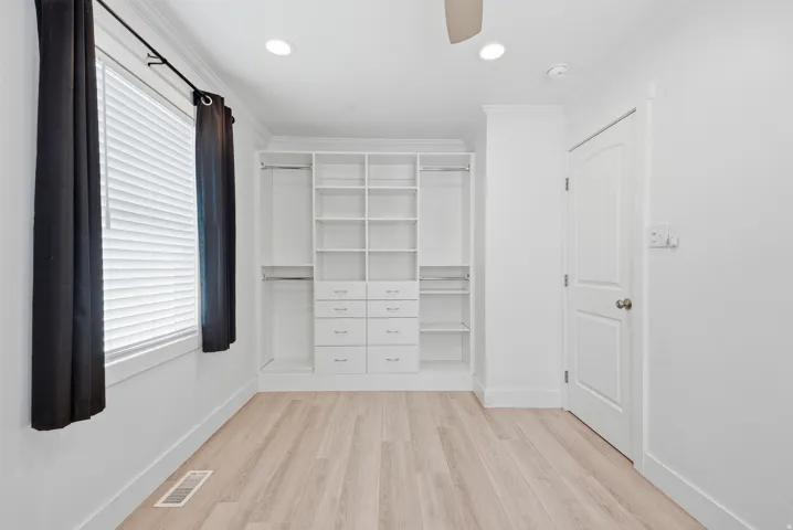 Light wood-finish flooring, built-in closet organization system with shelving and drawers, recessed ceiling lighting, and a ceiling fan