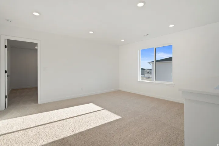 Bright, open bedroom with recessed ceiling lighting, large picture window framed in black, neutral carpeting, and access to an adjacent room via glass-panel door
