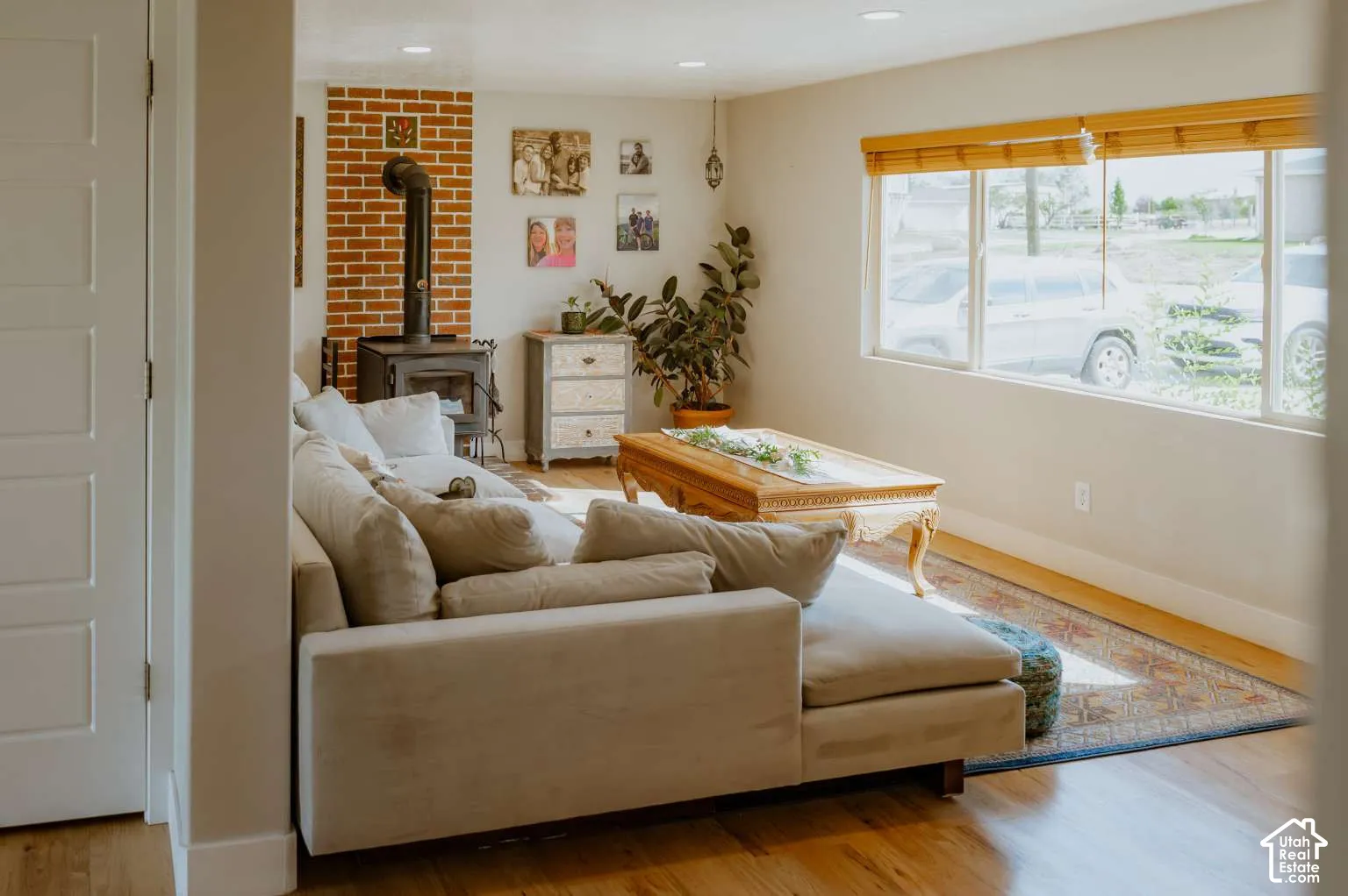 Living space featuring wood-finish flooring, a red brick chimney breast, a cast iron wood-burning stove, and a large horizontal window with natural wood-tone blinds