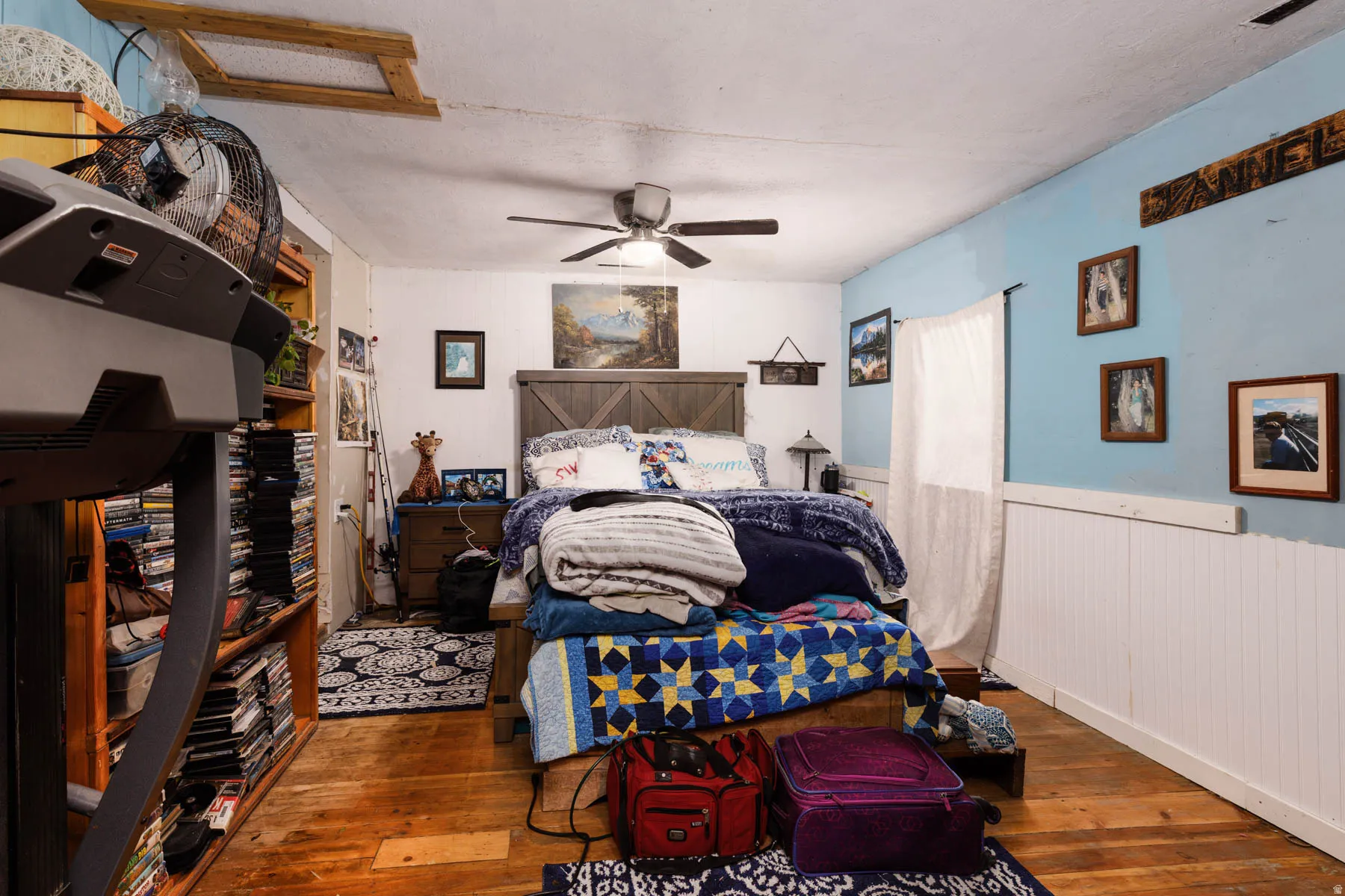 Hardwood flooring, painted blue walls with white wainscoting, wood-paneled bed frame, and ceiling fan with integrated lighting