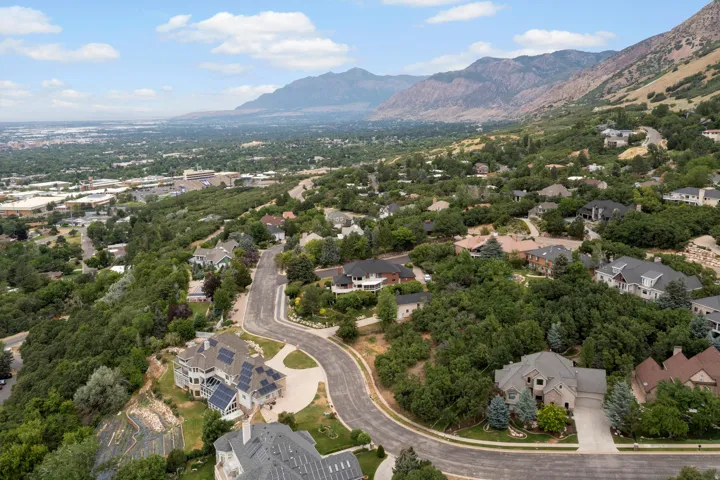 Aerial perspective showcasing this hillside residence nestled within mature landscaping, featuring a curved driveway approach, multi-level architecture, and panoramic vistas of the valley and mountain range beyond