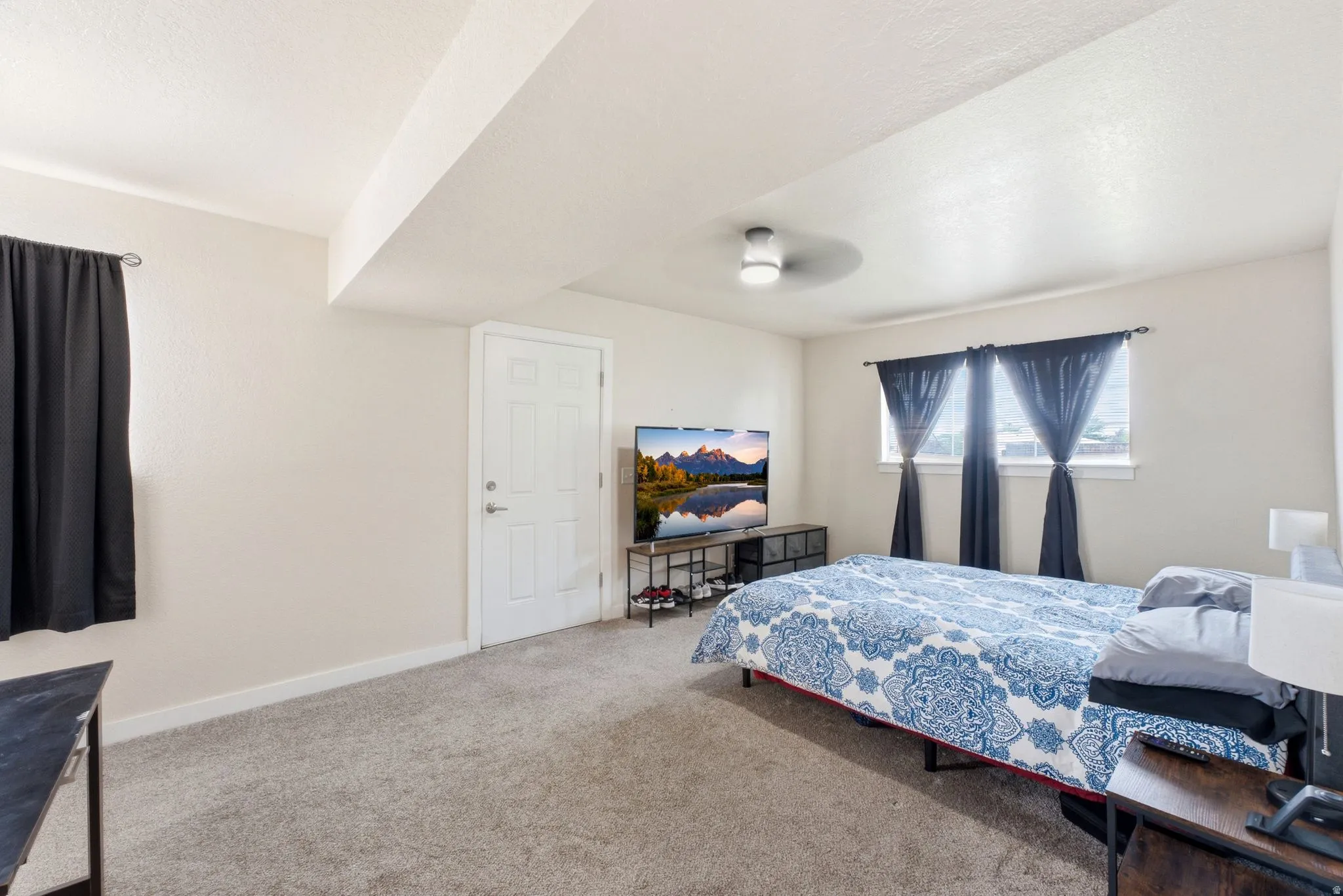 Spacious primary bedroom with carpet flooring, ceiling fan, and abundant natural light from dual windows dressed in navy curtains