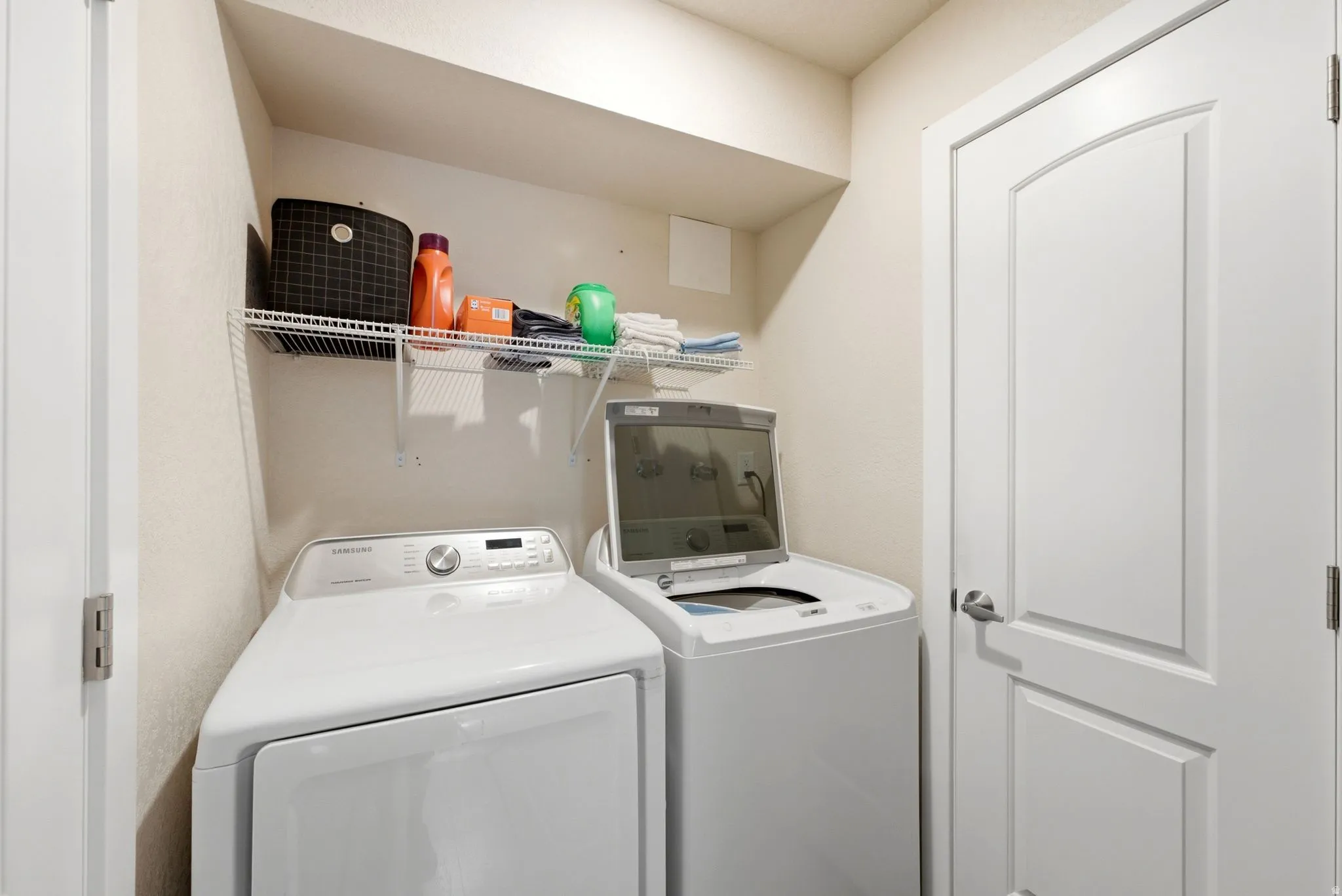 Dedicated laundry room featuring a front-load dryer and washer with stacked configuration, overhead shelving with storage baskets, and a finished interior with clean white cabinetry and neutral finishes