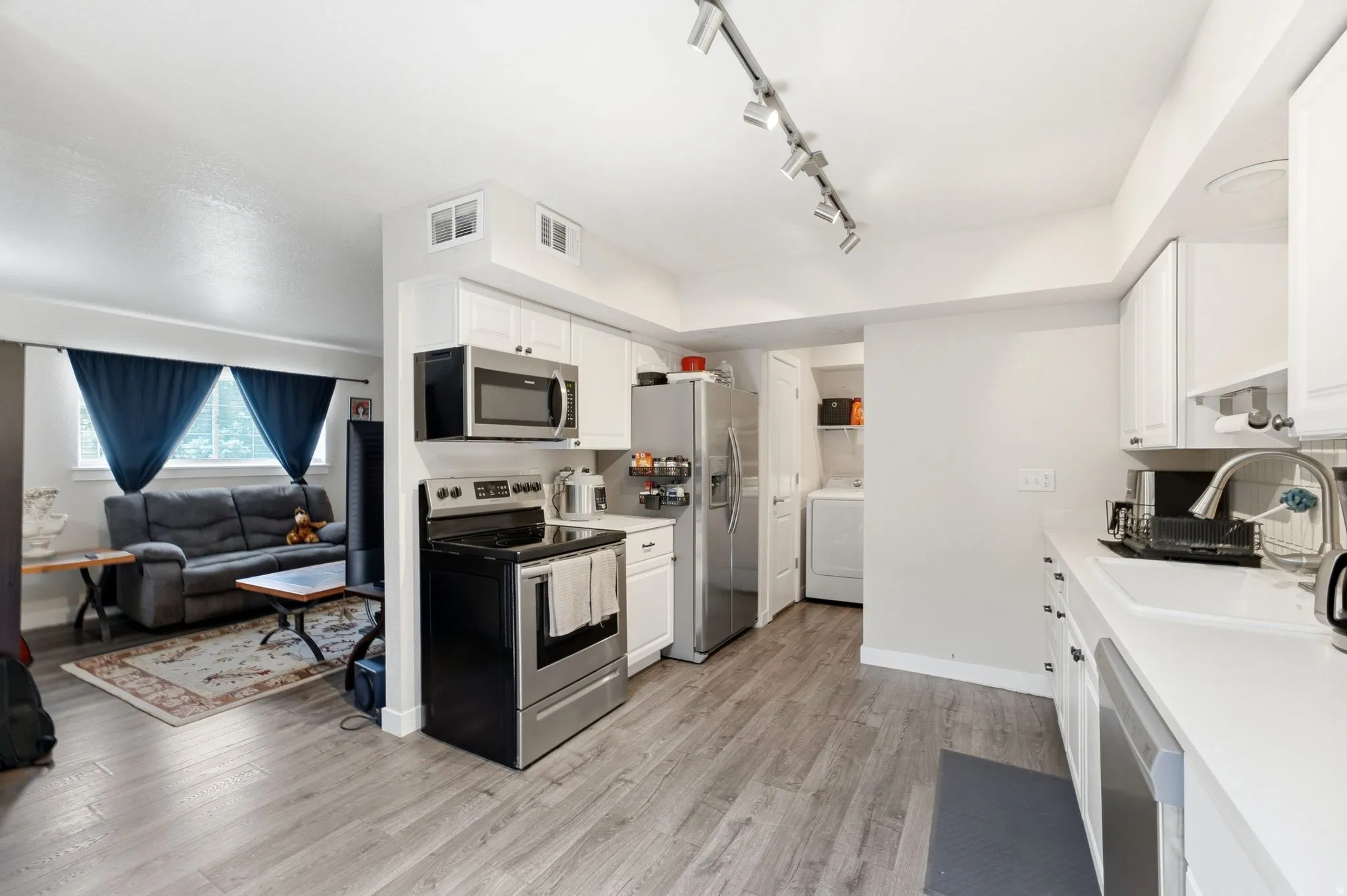 Open-concept kitchen and living area featuring stainless steel appliances, white cabinetry, wood-finish flooring throughout, and track lighting