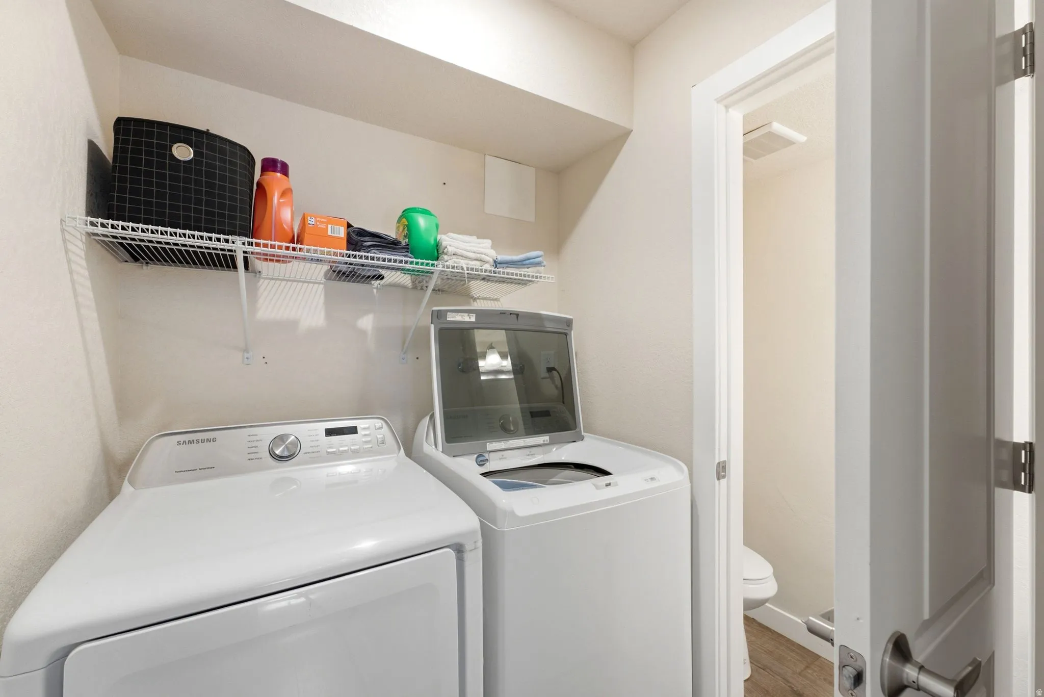 Dedicated laundry room featuring side-by-side washer and dryer with stainless steel finishes, wire shelving with organized storage, wood-finish flooring, and direct access to the home's main corridor