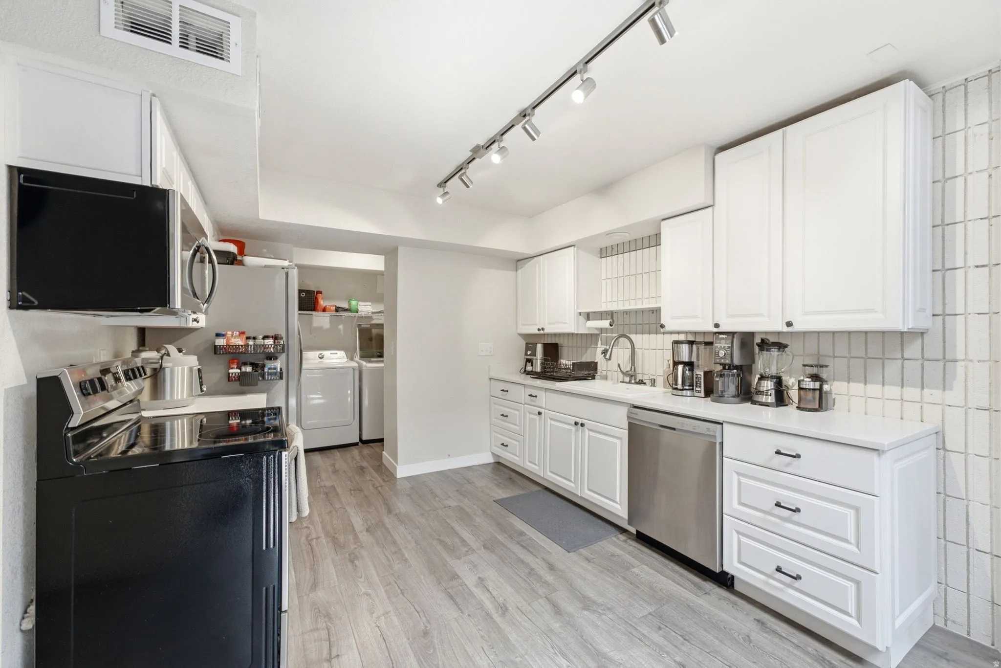 White cabinetry with dark hardware, stainless steel dishwasher, track lighting, wood-finish flooring, and a tiled backsplash