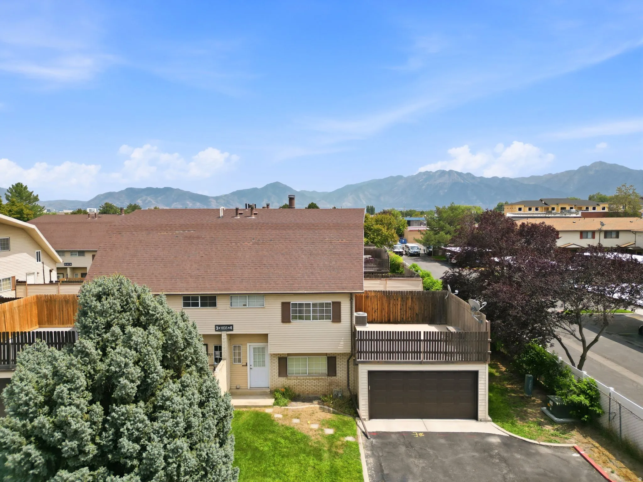 Attached garage with a private driveway, upper-level deck with slatted wood railing, and a mature evergreen tree