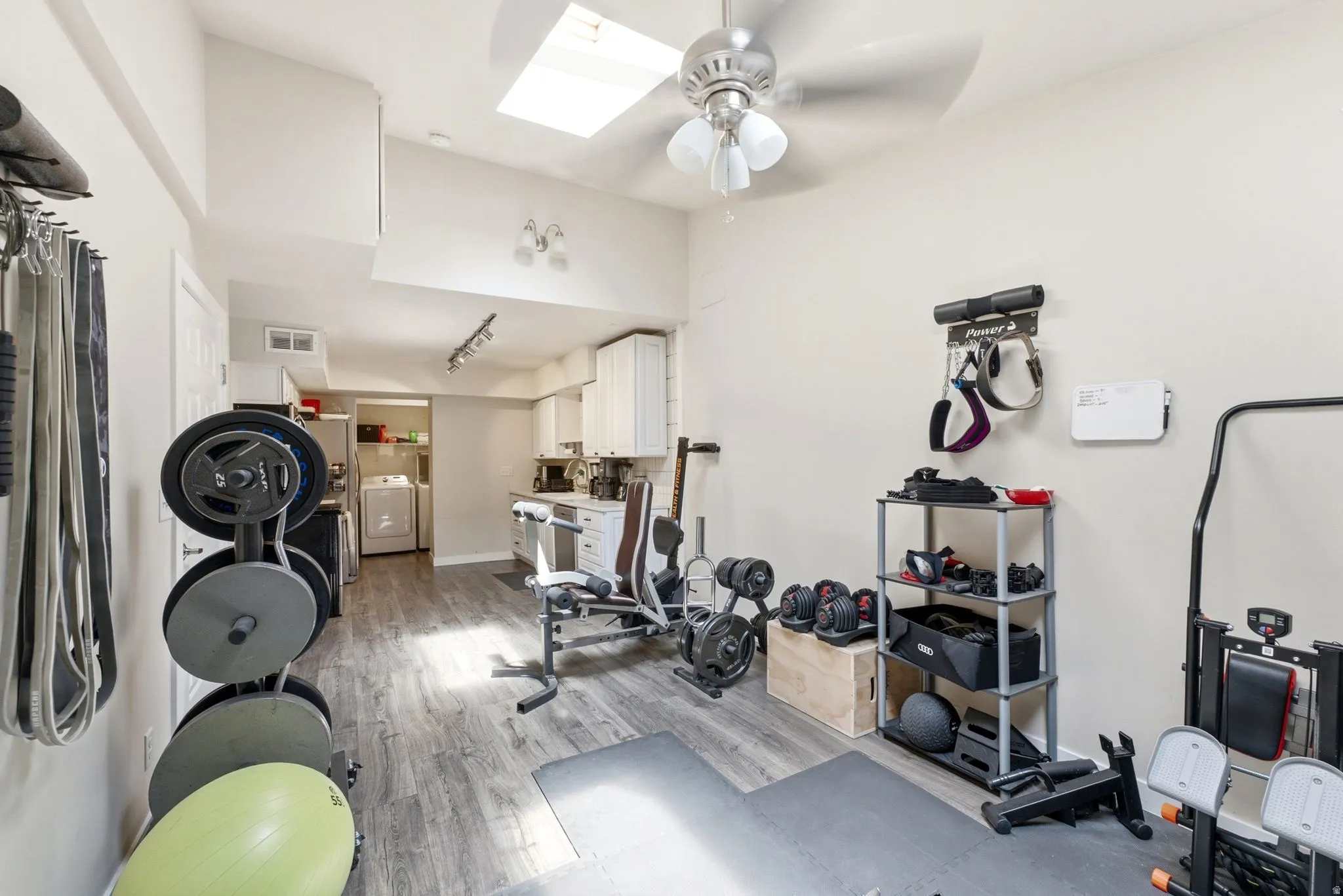 Dedicated home fitness studio featuring wood-finish flooring, ceiling fan with integrated lighting, wall-mounted weight plate storage, and ample space for cardio and strength training equipment