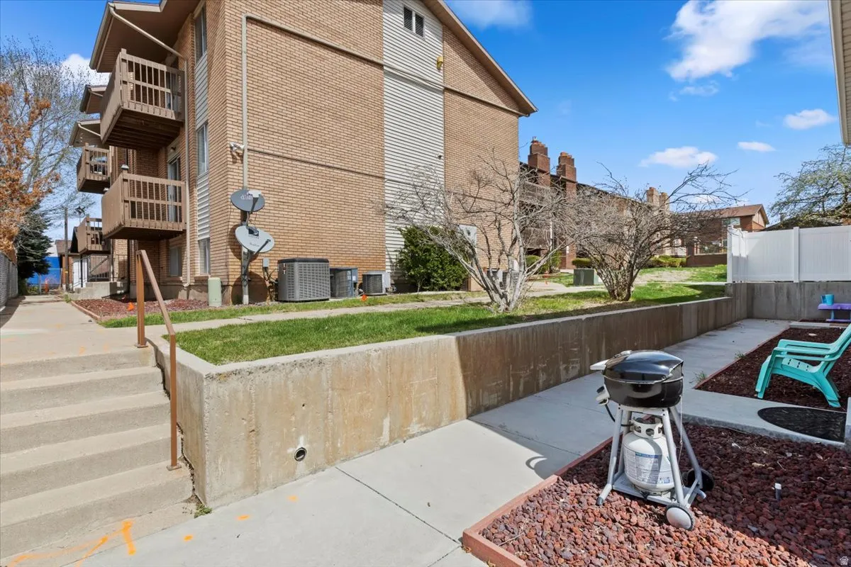 Brick multi-unit residential building with exterior staircase access, concrete patio area with mature landscaping, and dedicated outdoor space featuring mulched garden beds and metal fencing