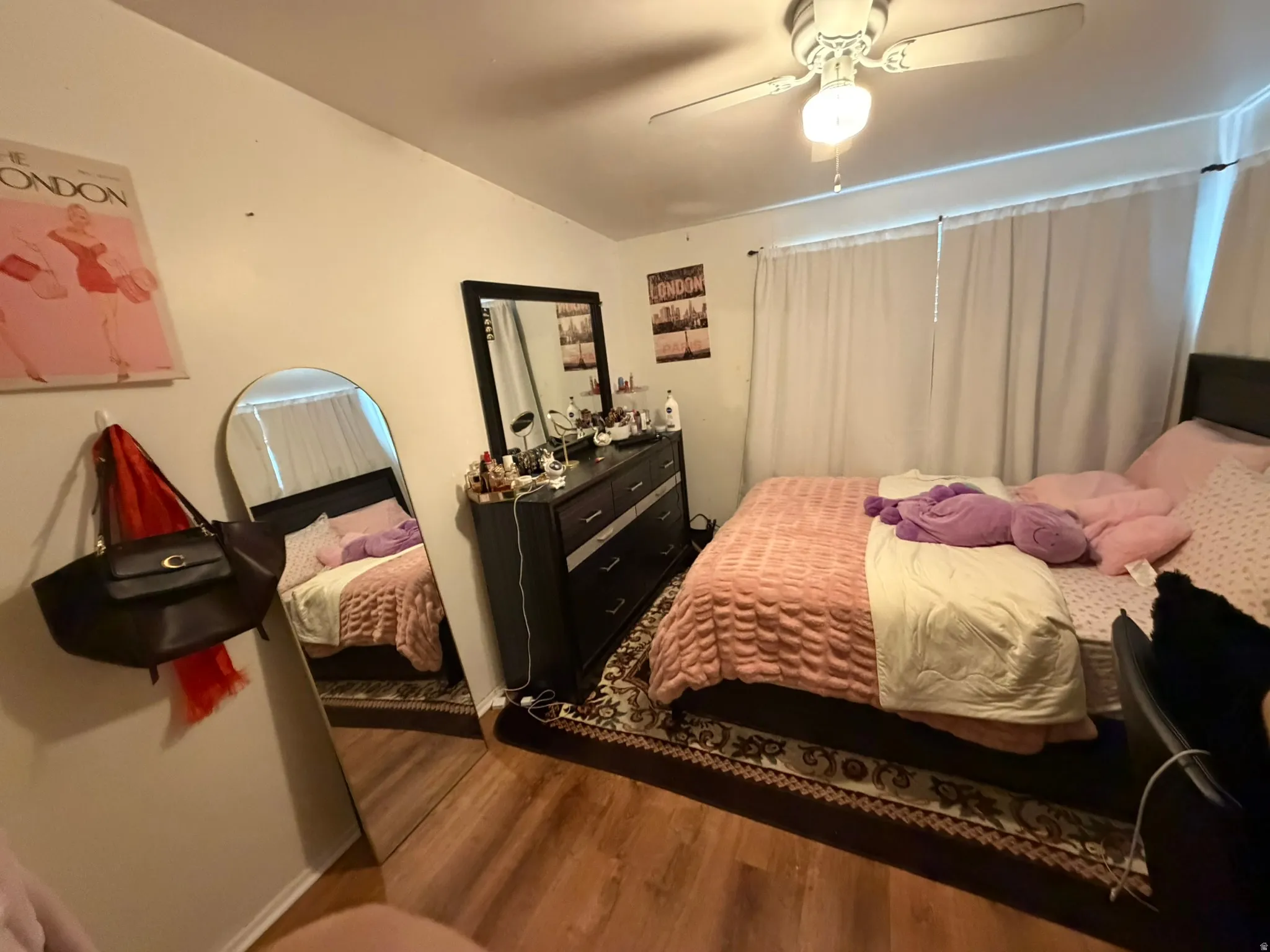 Wood-finish flooring, ceiling fan with integrated lighting, window with white privacy curtains, and wall-mounted hooks