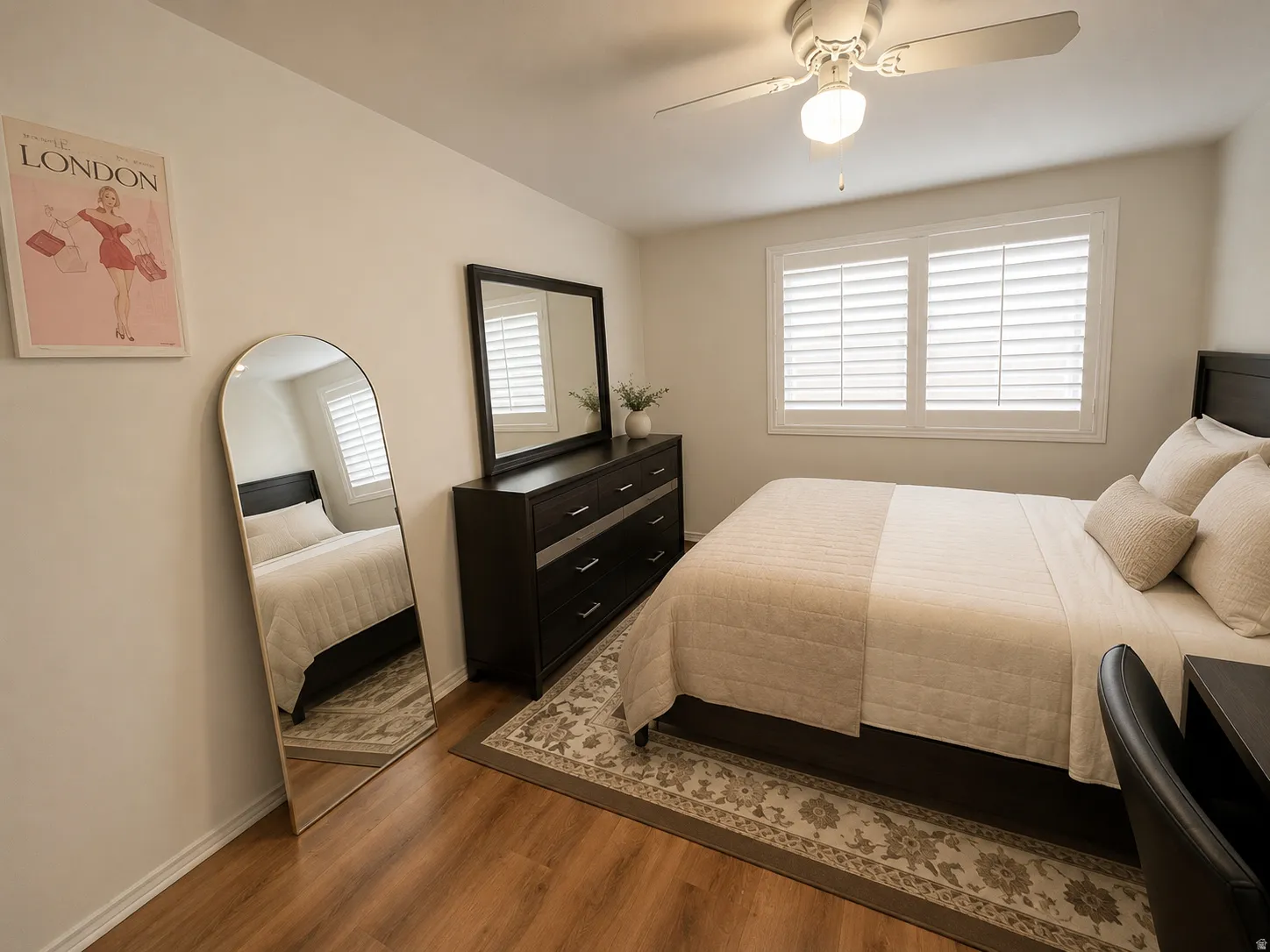 Wood-finish flooring, white walls, window with plantation shutters, ceiling fan with integrated lighting, and a dark wood-finish dresser with a mirror