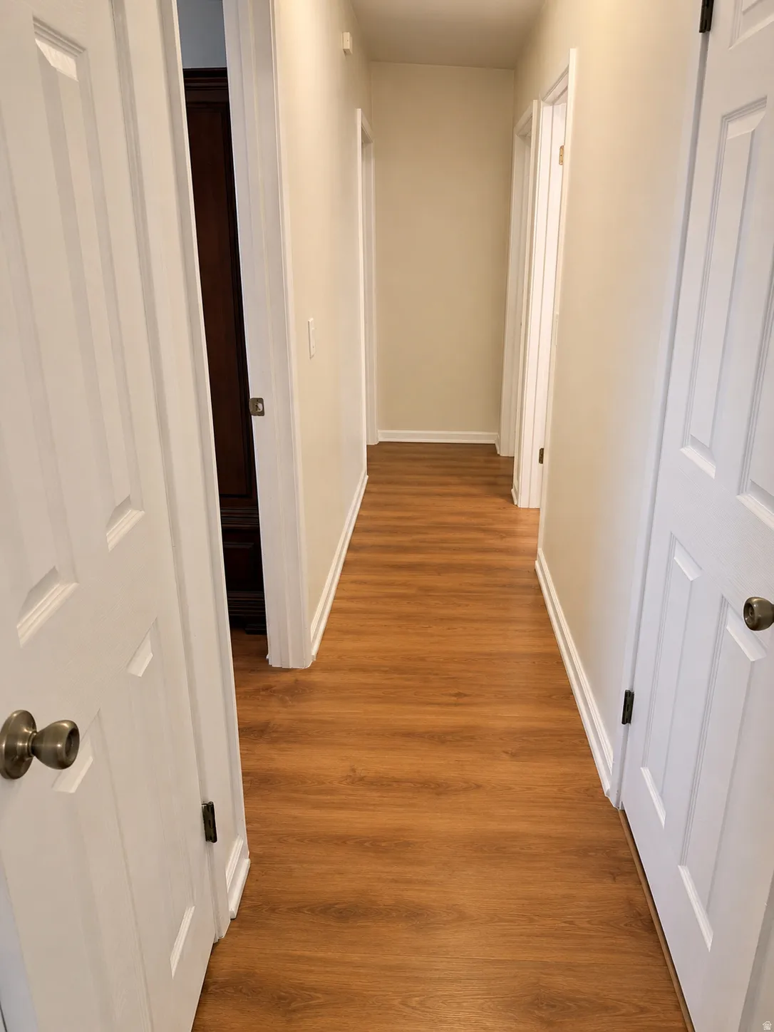 Hallway featuring warm wood-finish flooring, white six-panel doors with brushed nickel hardware, and clean white walls with crisp trim detailing throughout