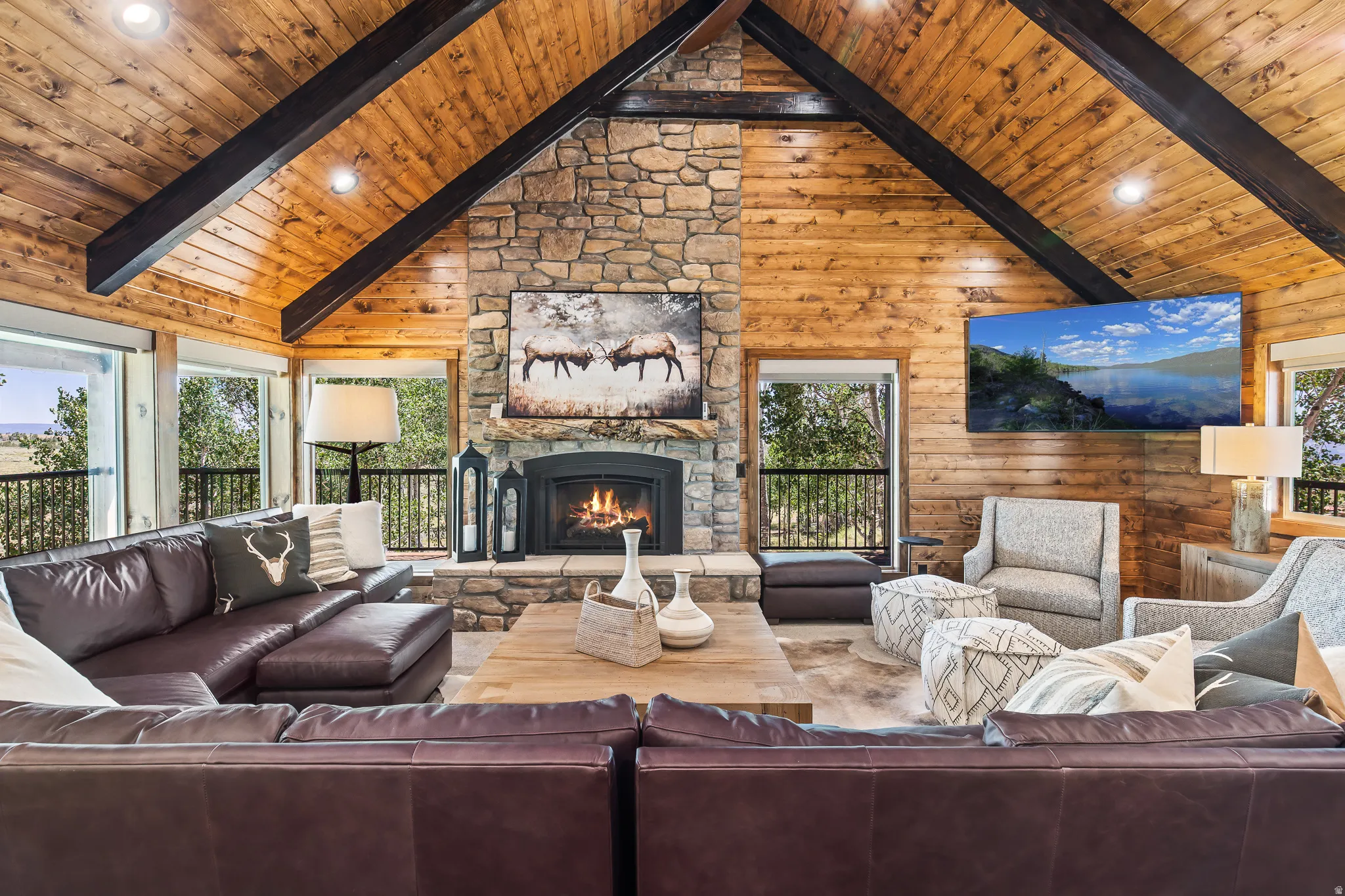 Vaulted wood-paneled ceiling with exposed beams, stone-clad fireplace, large windows, and a built-in entertainment area