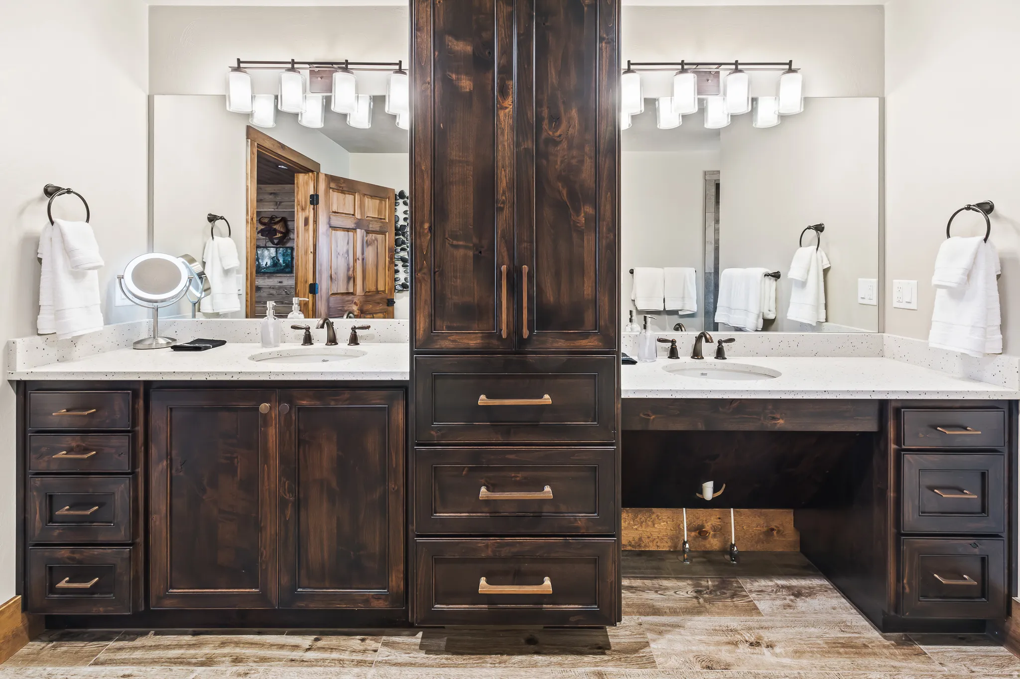 Dual vanity with light-toned countertops, integrated sinks, dark wood-finish cabinetry, and a central storage tower