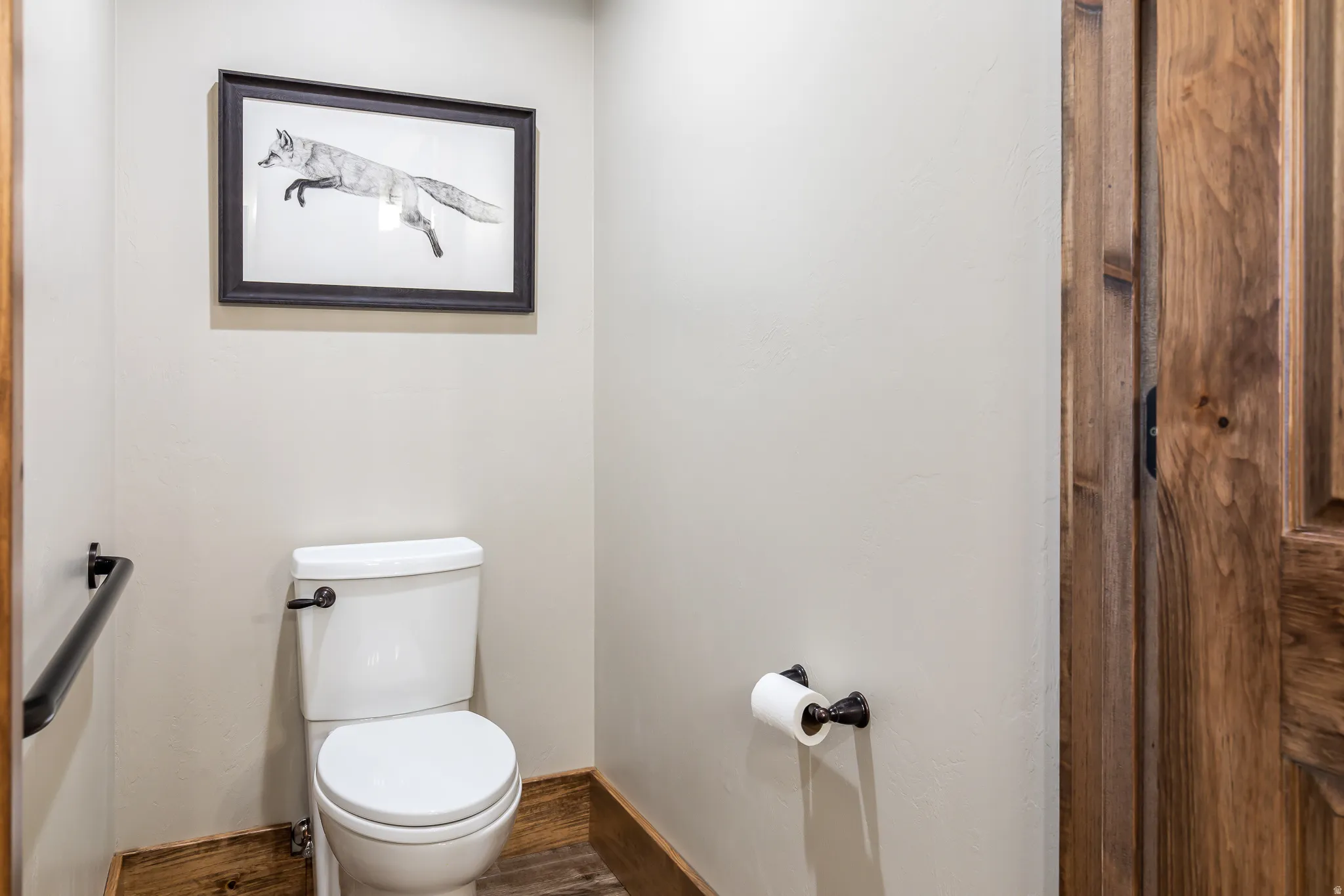 White toilet with dark hardware, wood-finish flooring, and a natural wood door