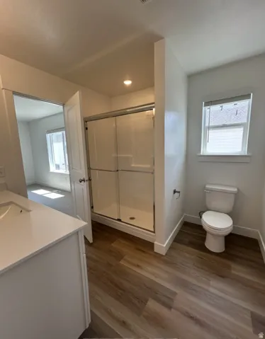 Wood-finish flooring, white vanity with integrated sink, glass-enclosed shower, standard toilet, and a window with blinds