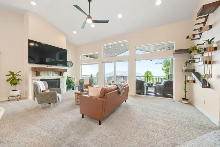 Vaulted ceilings, a stone-surround fireplace with wood mantel, recessed lighting, and expansive glass doors leading to an outdoor balcony