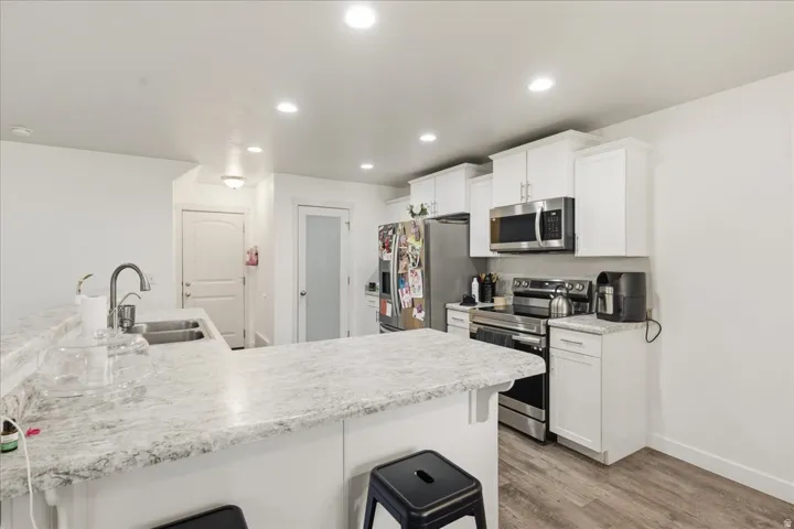 Kitchen featuring an L-shaped island with an undermount double basin sink, white cabinetry, stainless steel appliances, recessed lighting, and wood-finish flooring