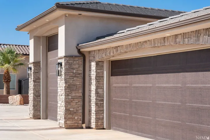 Stone veneer pillars, stucco exterior, textured garage doors, recessed soffit lighting, and dark tile roofing