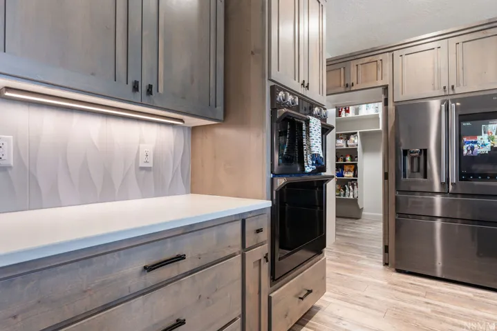 Light-toned wood cabinetry with dark hardware, white countertops, textured backsplash with under-cabinet lighting, stainless steel built-in appliances, and a walk-in pantry