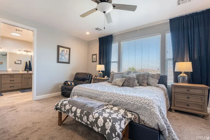 Recessed lighting and ceiling fan, large windows with blinds, plush carpeting, and an ensuite bathroom featuring a dual vanity with wood-finish cabinetry