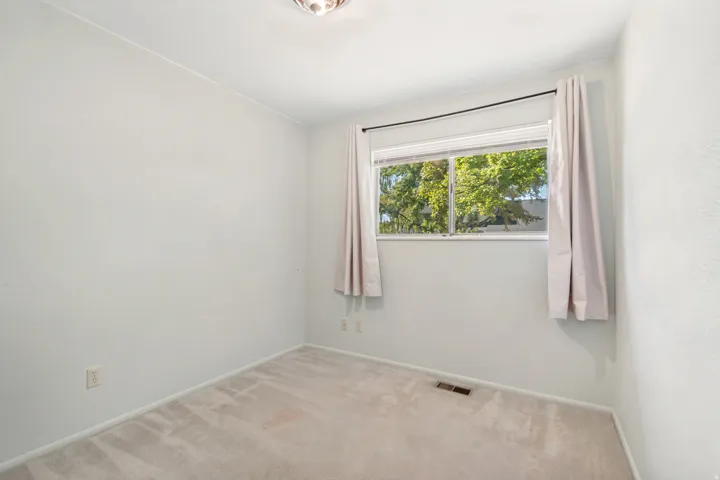 Bright secondary bedroom with large picture window framed by soft natural light, neutral carpet flooring, and fresh white walls