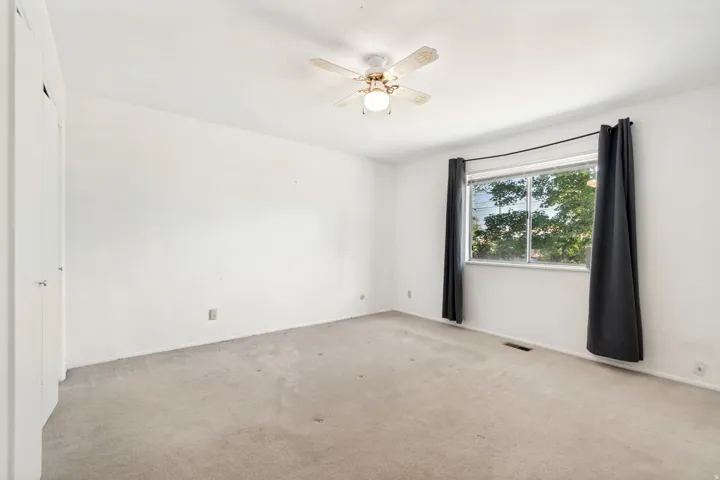 Spacious bedroom with neutral carpet flooring, ceiling fan with integrated lighting fixture, and large window with black-framed glass panes providing natural light