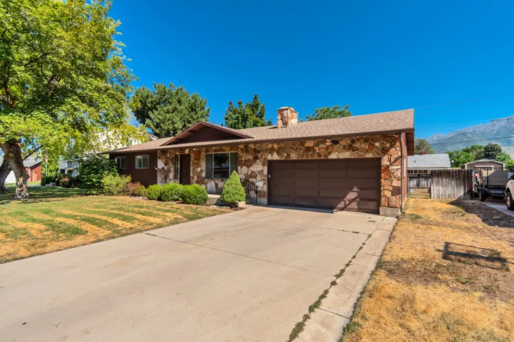 Charming brick ranch home featuring a wide driveway with attached two-car garage, mature landscaping with established trees, manicured lawn, and mountain backdrop