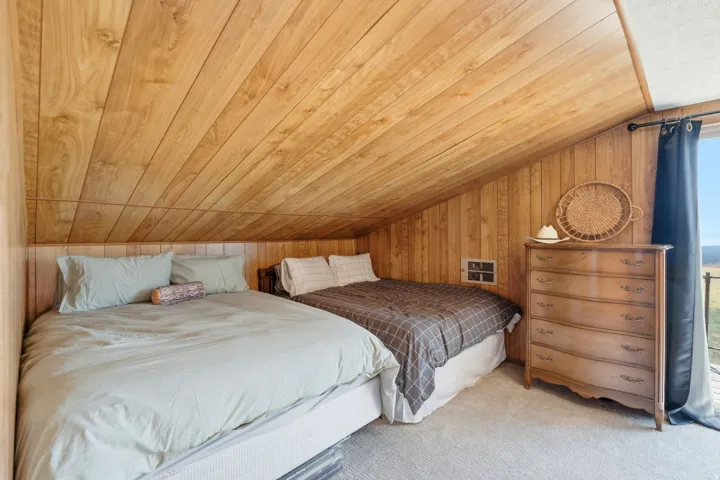 Cozy bedroom retreat featuring vaulted wood-plank ceilings, two queen beds with coordinating linens, wood-finish dresser with ample storage, and large windows with natural light and panoramic outlooks