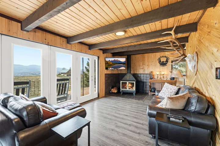 Open-concept living space featuring exposed wooden beam ceiling with recessed lighting, wood-finish flooring throughout, and expansive multi-panel glass doors opening to a covered deck with mountain vistas