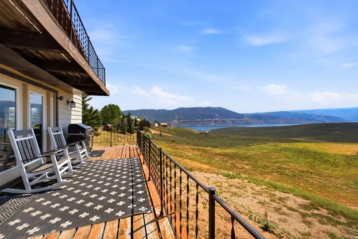 Expansive composite deck with checkered patterned flooring and metal railing overlooking rolling hillside terrain, distant water features, and mountain backdrop