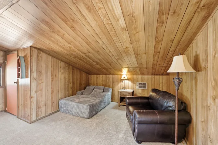 Rustic wood-paneled bedroom featuring cathedral ceiling with exposed beam details, carpeted flooring, built-in bedside lighting, and ample room for furnishings and storage