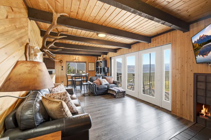 Open-concept living space featuring exposed wooden beam ceiling with recessed lighting, wood-finish flooring throughout, and extensive multi-panel glass doors and windows framing panoramic landscape vistas