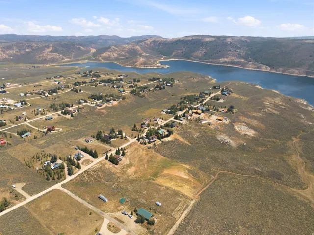 Expansive aerial perspective showcasing a residential community nestled within rolling terrain, featuring multiple homes with established landscaping, paved driveways, and direct access to a pristine blue-water lake with dramatic cliff-lined shoreline