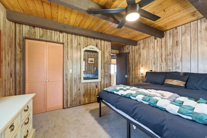 Bedroom featuring warm wood-paneled walls and wood-finish ceiling with exposed beams, ceiling fan with integrated lighting, carpeted flooring, and a wood dresser with multiple drawers