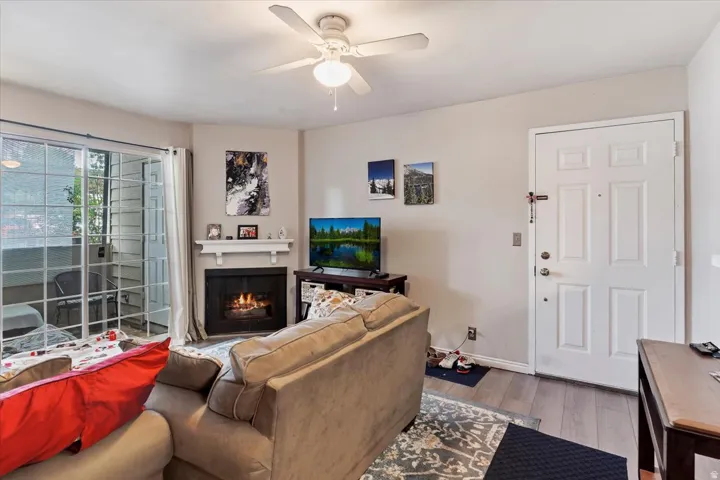 Living area with wood-finish flooring, a ceiling fan, and a fireplace featuring a white mantel