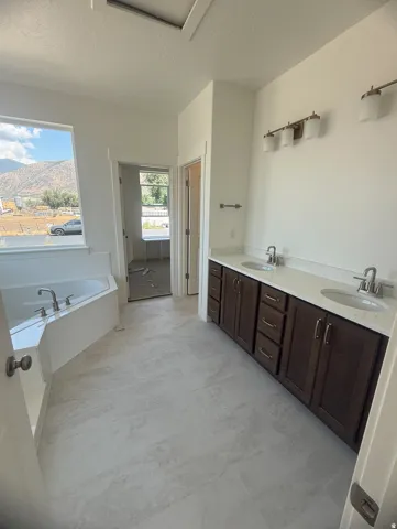 Large format tile flooring, built-in corner soaking tub, dual vanity with undermount sinks, and wall-mounted light fixtures