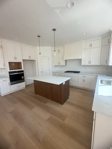 Contemporary kitchen featuring white shaker-style cabinetry, a two-tier island with wood-finish base and quartz countertop, stainless steel appliances including dual wall ovens and gas cooktop, wood-finish flooring throughout, and pendant lighting suspend