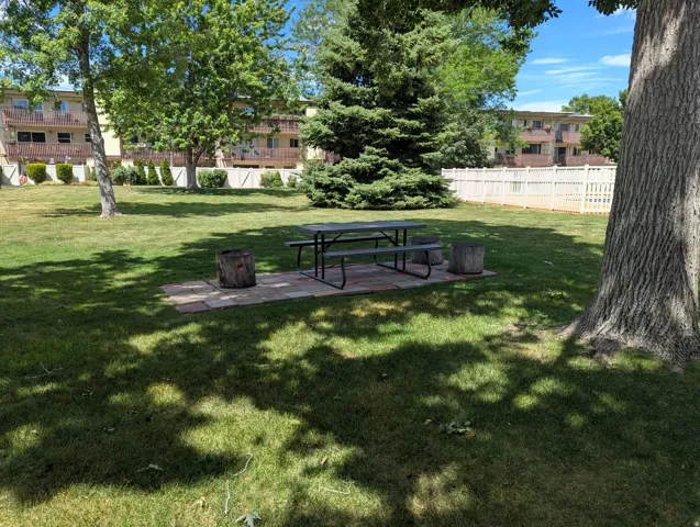 Grassed area with mature trees, a picnic table on a paver patio, and a white privacy fence