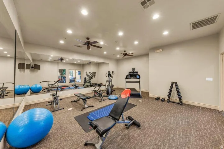 State-of-the-art fitness facility featuring multiple ceiling fans with recessed lighting throughout, mirrored wall systems for form feedback, durable textured flooring, and an array of exercise equipment including cardio machines, weight benches, and stab
